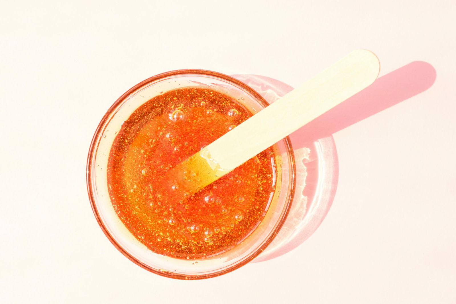 Shugaring paste in a glass bowl on a pink background. Hair removal, epilation, wooden spatula for sugaring paste | Live That Glow how long does a Brazilian wax last