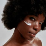 Close up portrait of young african american female model with afro hair looking at camera while posing with cream applied on her face isolated over gray background. Skin care concept