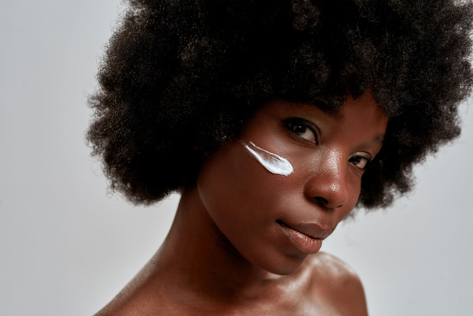 Close up portrait of young african american female model with afro hair looking at camera while posing with cream applied on her face isolated over gray background. Skin care concept