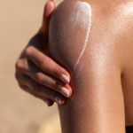 Young woman applying sun cream or sunscreen on her tanned shoulder to protect her skin from the sun. Shot on a sunny day with blurry sand in the background body