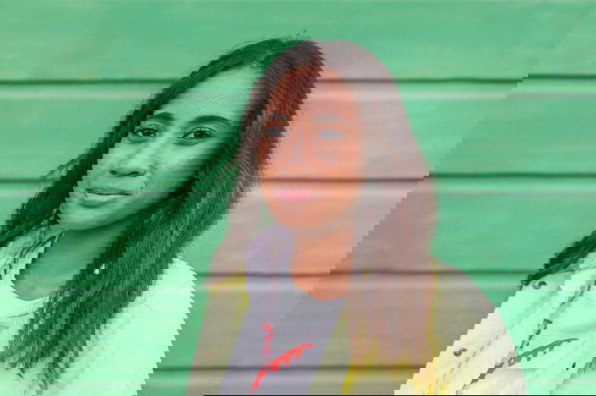 Young Pacific Islander female with long hair and acne looking at camera while standing against green cottage wall in Sonoma County, California